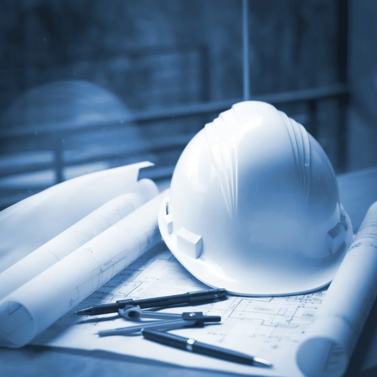 Casco de seguridad blanco sobre planos arquitectónicos enrollados, en tono azul, representando la gestión de proyectos de construcción